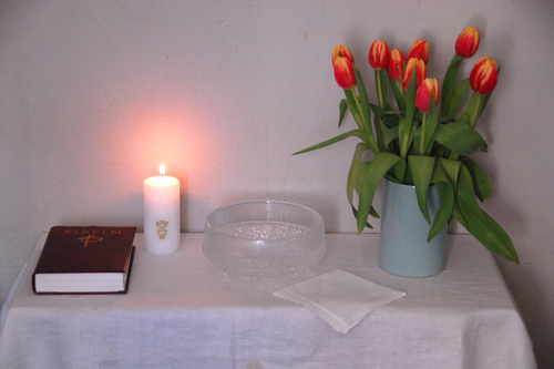 kastepöytä, jonka päällä on palava kynttilä, Raamattu, kastemalja, liina ja kukkia (tulppaaneita).